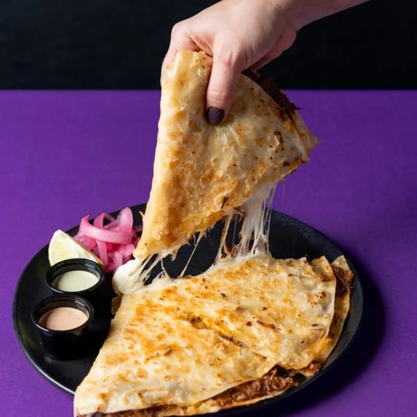 A hand lifts a cheesy quesadilla slice, stretching melted cheese, served with sauces and pickled onions.