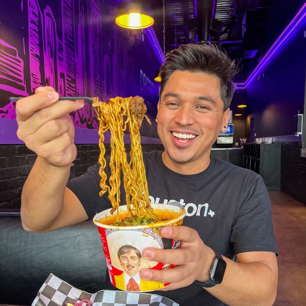 Man smiling, holding up a large serving of ramen noodles with chopsticks from a cup.