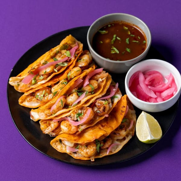 Shrimp tacos with pickled red onions, cilantro, a side of consommé, and lime on a black plate.