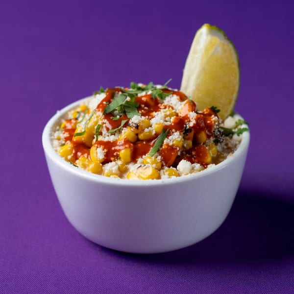 Elote in a white bowl, topped with red sauce, cheese, cilantro, and a lime wedge.