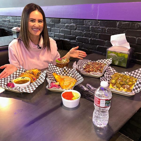 A smiling woman sits at a table with four trays of food, including tacos and nachos.