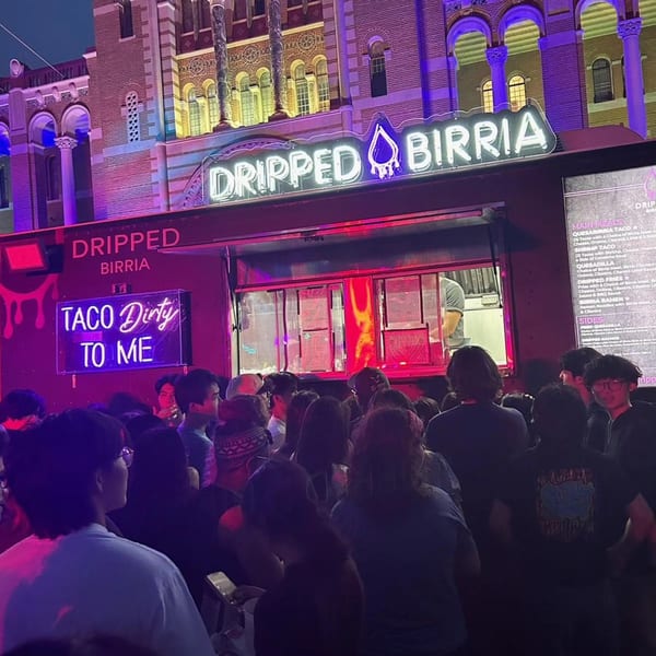 A crowd of people gathers around the "Dripped Birria" food truck at night, illuminated by neon signs.