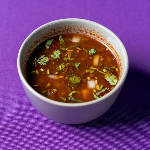 A white bowl of rich red soup with cilantro and diced onions on a purple tablecloth.