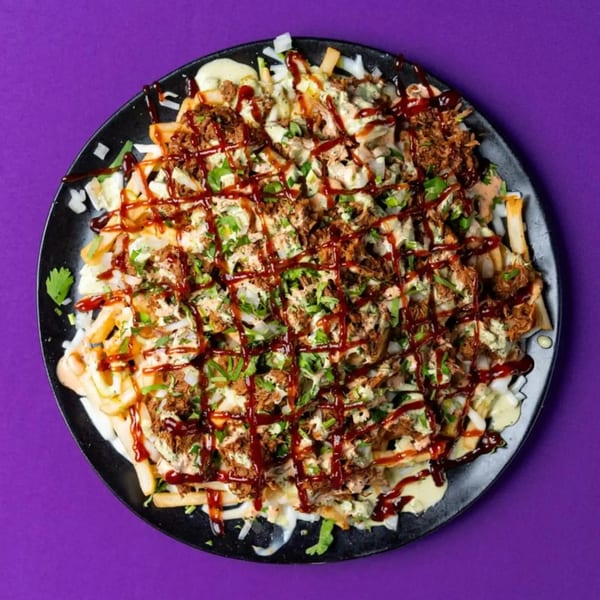 Loaded fries with pulled pork, white sauce, barbecue drizzle, and fresh cilantro on a black plate.
