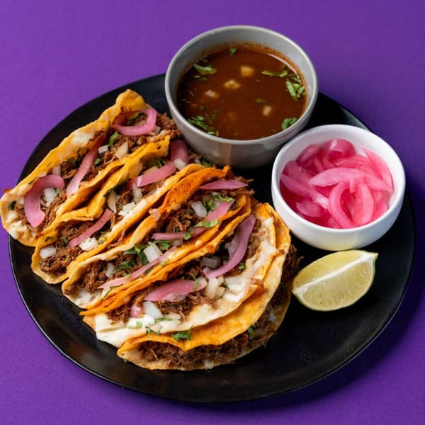 Birria tacos with consommé, pickled onions, and a lime wedge on a black plate.