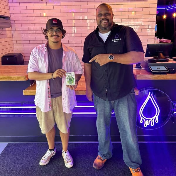 Two men, one holding a gift card, stand smiling in front of a counter with a neon sign.
