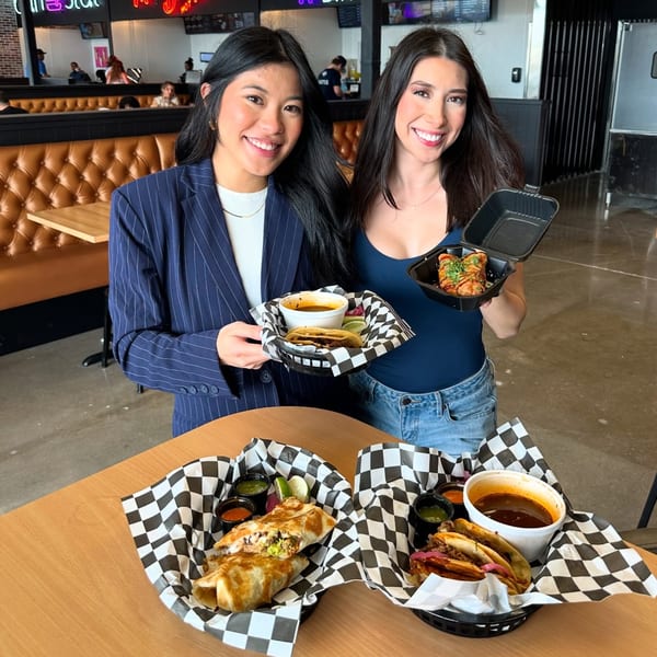 Two smiling women holding baskets of tacos and burritos with dipping sauces in a restaurant.