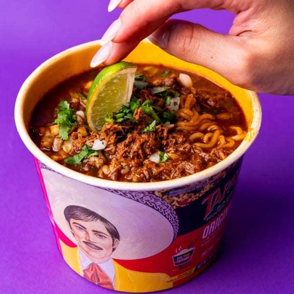 A hand squeezes a lime wedge over a cup of birria ramen with cilantro and onions.