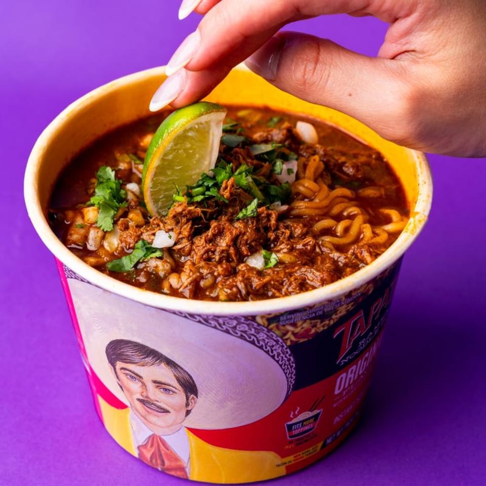 A hand squeezes a lime wedge over a cup of birria ramen with cilantro and onions.