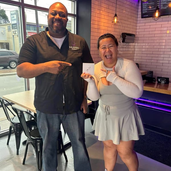 Two smiling people, a man and a woman, pointing at a small white box in a restaurant.